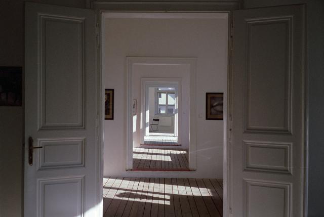 A series of open doors leading through multiple rooms, with the last room containing a single chair and a window.
