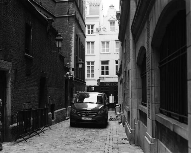 A black and white photo of a car parked in the narrow, walled-off space between two old buildings.