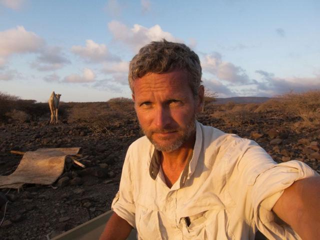 Selfie photo of a man on a rocky hillside with a light beard, a loose-fitting shirt, and a camel in the background.