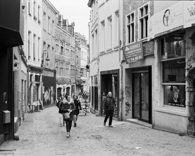 A black and white photo of a cobblestone paved street with old buildings on each side and people walking along.
