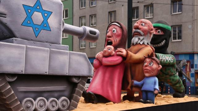 Photo from carnival procession in Cologne 2024. It shows an Israeli tank pointing its gun to a Palestinian family behind which a grimly laughing male in green army cloth with a black hat holds them as human shield. The picture doesn't show any of the IDF brutality or war crimes that happened in Gaza in 2023 and 2024