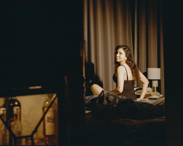 A beautiful woman with black hair, dressed in a bra and a corset, is sitting on the bed and looking over her shoulder, framed by the doorframe.