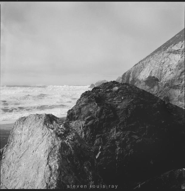 a square black and white photo, a beach scene. In the foreground, two large boulders. The one on the left is light gray, the one to it's right is very dark. There is a great deal of rough surface visible on both of them. To the right, a hill descends toward the ocean. The waves can be seen in the middle distance, row upon row of them approaching the shore. The sky is mostly cloudy.