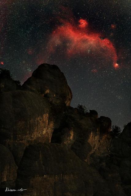 A red nebula in a dark starry sky is seen above a rocky peak. The nebula appears similar to a flying bird.