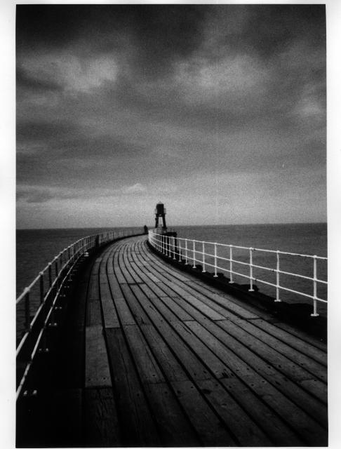 A black and white print of  a wooden pier leading to a lighthouse 