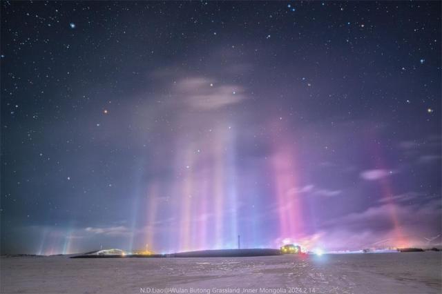 Numerous thin pillars of light connect a landscape filled with snow to a star filled sky. The Big Dipper can be seen through the colorful pillars.