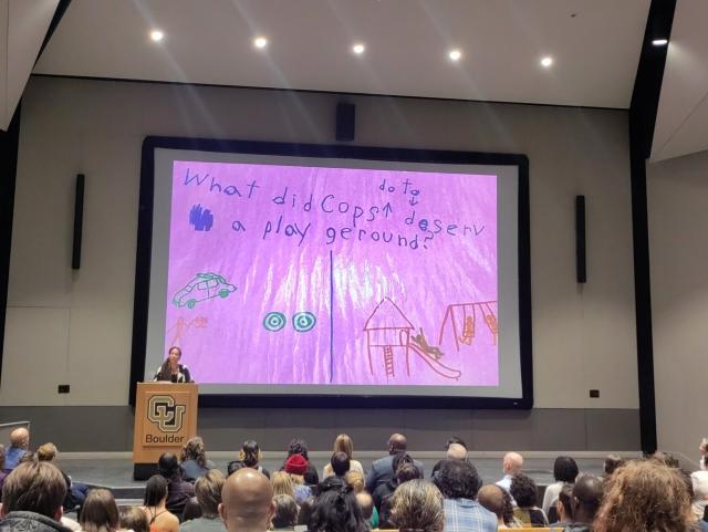 Lecturer at a podium in front of a slide of a child's art that asks "What did cops do to deserv a play geround?"