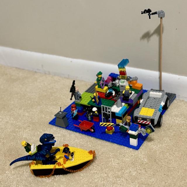 A colorful Lego scene depicting a velociraptor being taken toward an island of different warriors awaiting battle or waiting to greet it or something else?