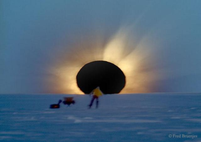 A field of snow leads up to a dark circle. Light rays eminate from this circle. In front, standing on the snow field is a person and to the left is a folding chair and a bag.