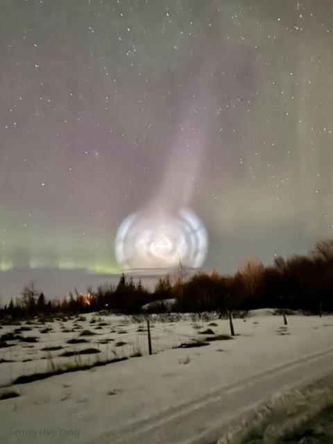 A field of snow is shown, lined with trees along the back. Above the horizon is an unusual white spiral cloud. Stars dot the background, and faint green and red aurora are also visible.