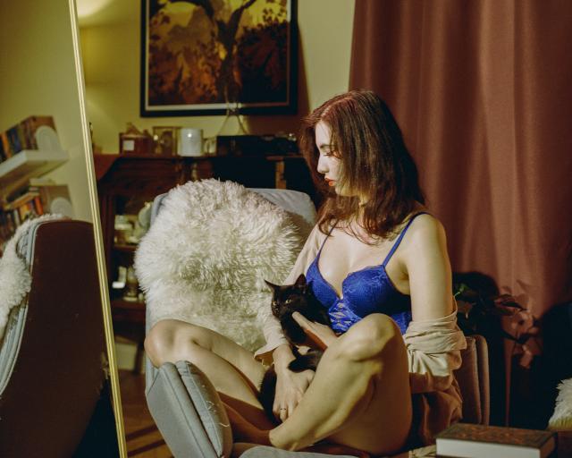 A beautiful woman with dark hair, wearing blue lingerie and a beige blazer, sitting cross-legged on a lounge chair while holding a black cat in her arms. The cat is giving you, the viewer, a side eye.