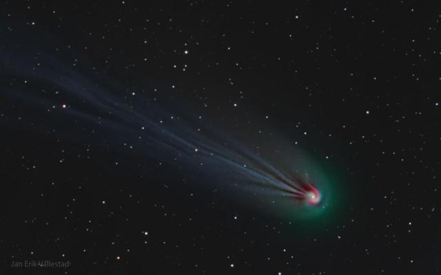 A comet is pictured with a really long and wavy ion tail. The front of the comet -- its coma -- appears to be a spiral. The coma is green, the tail is faint blue, and part of the swirl is red.