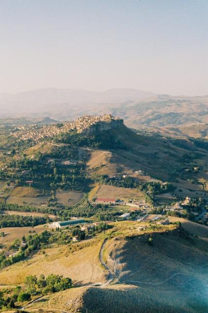 Photo of an aerial-like view of a city in the mountains. In the background there are mountains. Around the city there are trees, roads and fields.