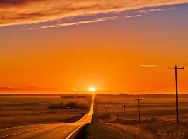 The sun sets in the distance at the horizon end of a long road over open country. The sunset is very orange, as is the surrounding sky. Telephone poles line the right side of the road.