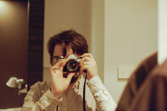 A white man with black hair, black rim glasses, and a ring on his finger is holding a Canon AE-1 Program camera, as seen from a mirror of a hotel room.