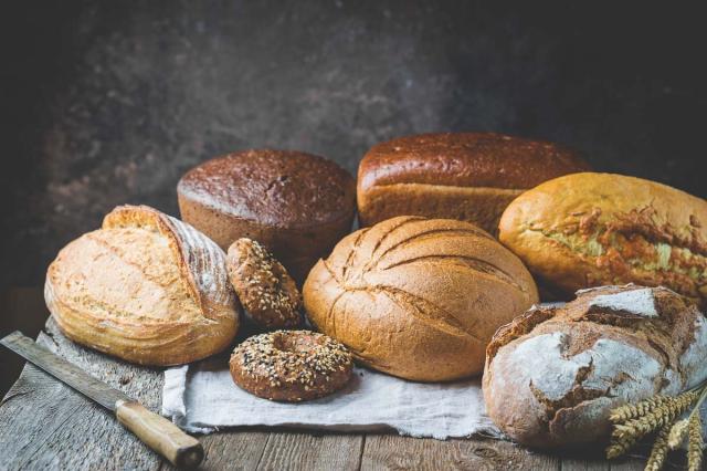 This is a photo of a large variety of German bread.

It shows various loaves, from light wheat to dark rye. There are two rolls with seeds, and breads that are round, oval and square. Some with a glossy top and some with a crunchy crust. Some floured and some plain.

The breads are on a wooden table with a white linen cloth underneath them. There is also a large rustic bread knife.