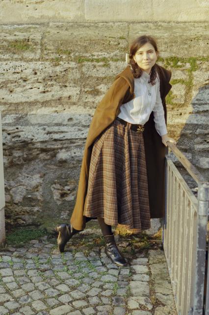 A beautiful woman with black hair, dressed in a white shirt, olive cap, tartan woolen skirt, a black belt with a metal bee for a buckle, French heel shoes, and a snake necklace, is playfully leaning on a metal guardrail.