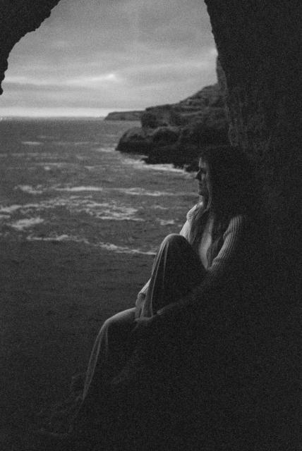 A woman sitting in a cave, she looks thoughtful, in the background are cliffs and the sea.