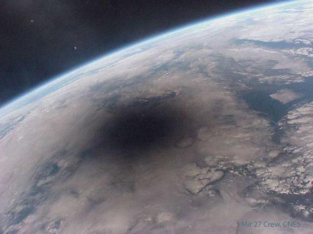 Part of the the Earth is pictured with blue seas and white clouds. On the upper left is a deep space dark background. On the Earth a large dark spot is apparent.