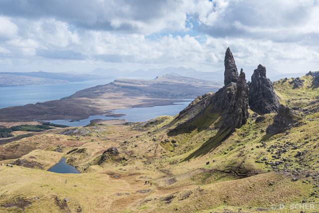 The Old Man of Storr, a 55-meter-high basalt pinnacle, on a sunny day with a few clouds, looking out to sea over an island and then beyond to shores and mountains. 
A small group of hikers can be seen at the bottom of the photo, giving a better idea of the scale.
