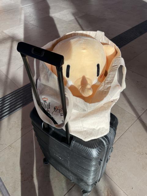 A plush Mastodon in a tote bag on top of a large suitcase at an airport. 
