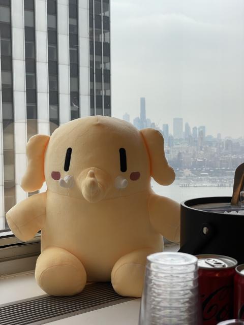 A stuffed Mastodon toy sitting in front of a window of a tall building with the skyline of New York visible in the background. 