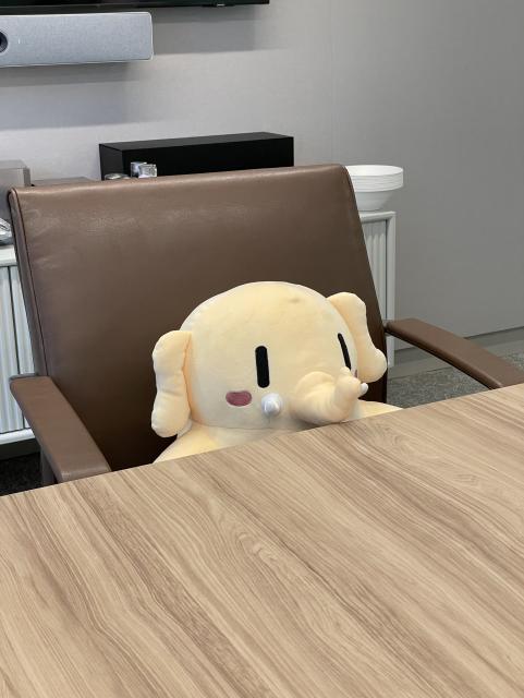 A stuffed Mastodon toy sitting in a large executive chair at a conference table. He is tall enough that his trunk is above the table. 