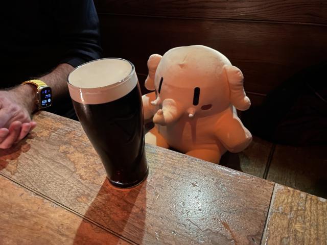 A stuffed Mastodon toy sitting at a wooden pub table in front of a glass of beer. 