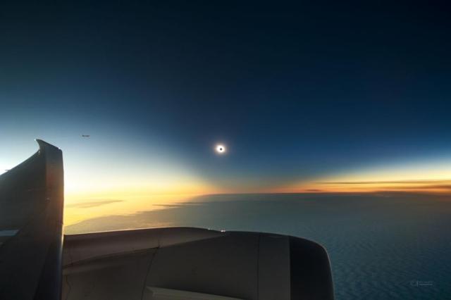 A totally eclipsed Sun is seen in the distance. Around the eclipse is a dark region dipping down from above. Below that are clouds and below that is the wing and engine of an airplane.