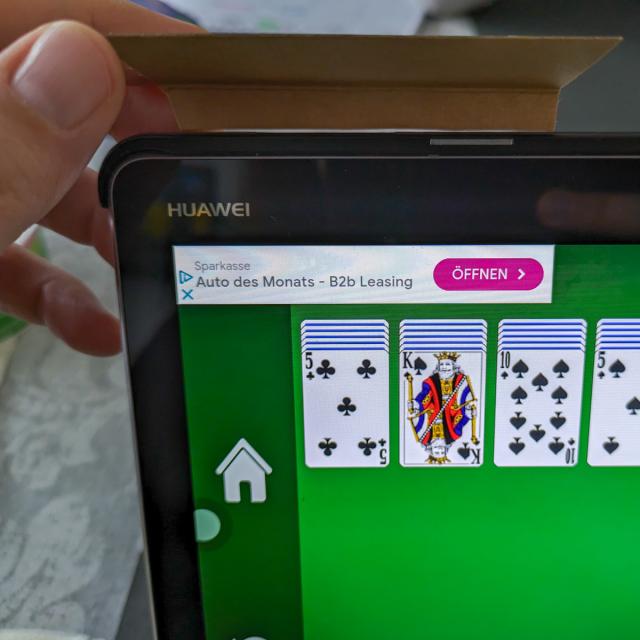 Close-up of the upper left corner of a Huawei tablet. The screen displays a card game with an ad banner in the top left corner. There's a hand holding a piece of cardboard taped to the corner of the tablet.