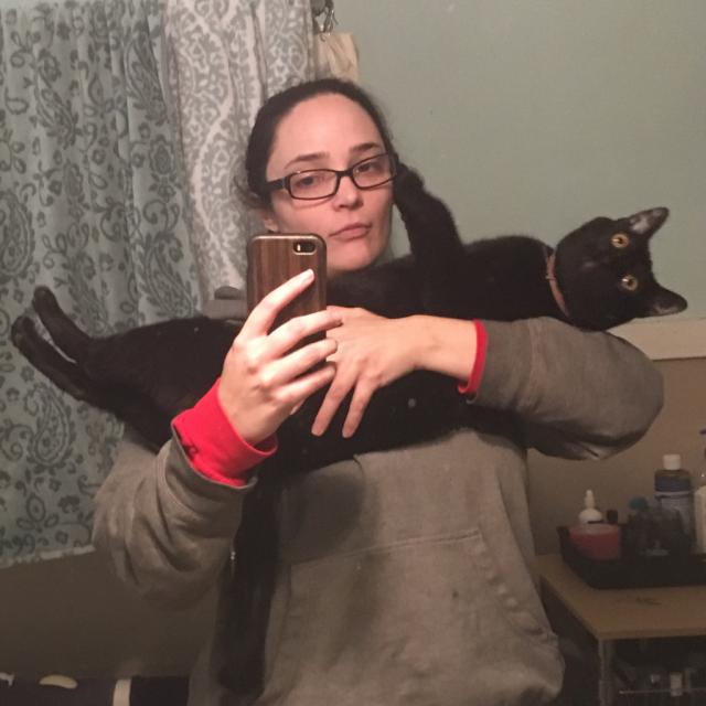 I’m taking a mirror selfie in a bathroom mirror. I’m trying to cradle my black cat Shadow in my arms but instead he’s holding himself stock straight with his legs straight out and he’s looking up at an angle with his eyes open incredibly wide and frozen. I have my hair pulled back and I’m wearing my old rectangular framed glasses and I’m pursing my lips to keep from laughing and ruining the shot. I cannot overstate how much he looks like bad taxidermy here. A living cat should not look this wooden and awkward. It also really showcases how huge he is, he’s inches longer than my shoulders are broad not including his leggies 