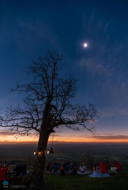 A totally eclipsed Sun is seen in the sky surrounded by a bright corona. In the foreground several people watch it near a large tree. To the right of the eclipsed Sun is the bright planet Venus, while the nearly-as- bright planet Jupiter is to the left.