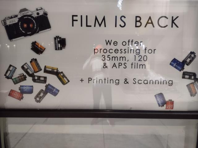 Photo: Large sign in window states:
"FILM IS BACK
We offer
processing for
35mm, 120
& APS film
+ Printing & Scanning"

The photographer's reflection can be seen in the window.
