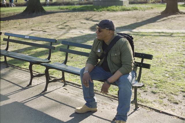 A man with a double pierced ear in a green jacket, black t-shirt, and jeans, wearing a cap and shades and a Rebel Alliance bracelet, sitting on a bench in a park.