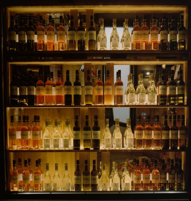 Das Foto zeigt ein Fenster einer Bar. Auf mehreren Ebenen sind Weinflaschen zu sehen, gefüllt mit weissem, roten und Rosewein. Die Flaschen sind beleuchtet, da es eine Nachtaufnahme ist. 
