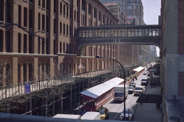 Two red brick buildings connected by a skybridge and a busy road underneath it. The sun is reflecting intensely off the roof of a bus stop.
