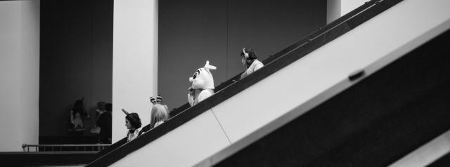 A black and white photo of four people on an escalator. The person in the middle of the photo is wearing an Easter Bunny costume. There are three columns in the background with some people in shadow between the first and second columns