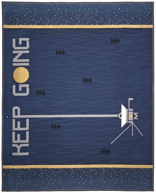 Voyager II themed quilt. The background is navy with wavy metallic thread quilting. Along the left side, from top to bottom, pieced letters read KEEP GOING with the “O” replaced by an embroidered Golden Record. In the lower right corner is a pieced image of the spacecraft. To the left of the “P” is embroidered an pale blue dot and the “P” itself has a long tail that reaches to the center of the spacecraft’s dish, like a communication between Voyager and Earth