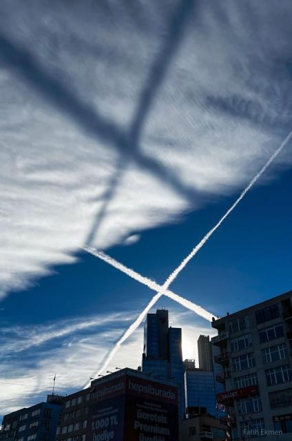 Two airplane contrails, crossing in an X, are shown across the middle of the image. They are bright white against a dark blue background. A high cloud deck is seen above the crossing, sunlit, contrails. A low Sun creates a dark shadow X on the high while clouds. A row of buildings runs across the lower part of the image.