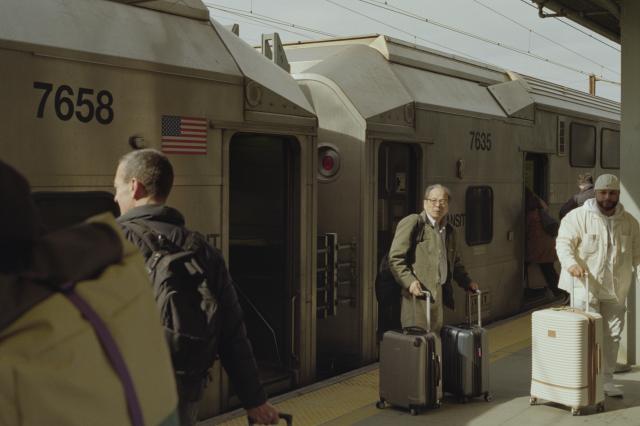 A bulky metal train with open doors standing on a platform. People are boarding. One man with a suitcase stands in front of the train, illuminated by bright sunlight.