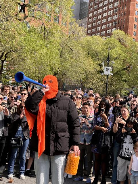 A man in an orange ski mask holding a giant jar of cheese balls addresses a crowd through a blue station horn.