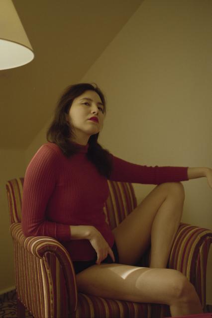 A woman with black hair and red lips, wearing a red turtleneck sweater, sitting in a relaxed pose on a colorful chair.