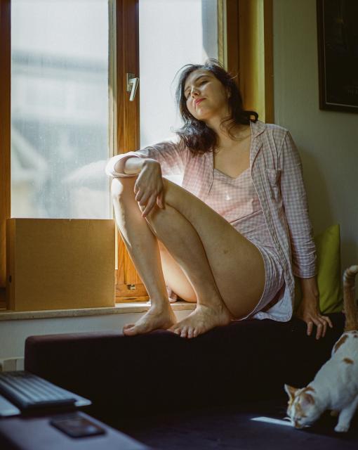 A woman with black hair dressed in a pink checkered pyjama sitting on the edge of the window, illuminated by bright beams of sunlight.