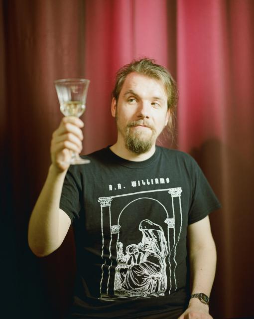 A man with green eyes, long blonde hair and blonde beard, wearing an A. A. Williams t-shirt, holding a crystal glass in front of a red curtain.