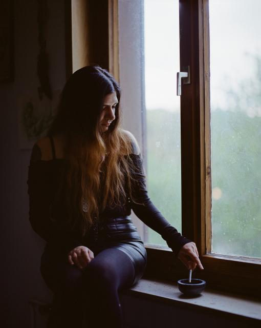 A woman with black hair that has a gradient towards the ends and a septum ring, wearing a large round silver pendant, black tights, a black faux leather skirt, and a black off-the-shoulder top, with tattoos peaking out from underneath the top on both arms, sitting on the windowsill and dipping a cigarette gently into the ashtray next to her.