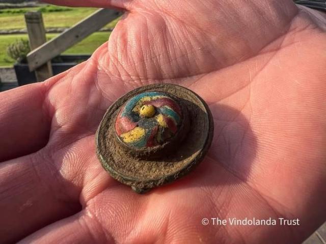 The brooch has a multi-colored molded glass inset mounted into the central setting, with red, blue and yellow stripes swirling out from a yellow nipple. 
It has a low wall outlining a round central setting (multi-colored) and a ridge around the outer edge. It is made of brass and would originally have been gilded. 