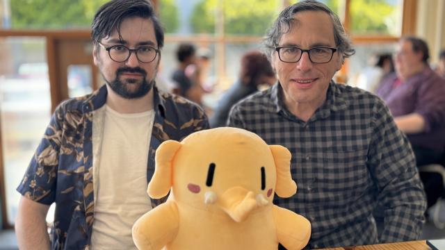 Two people are sitting at a table with a yellow plush elephant toy between them. The background shows an indoor setting with other people.