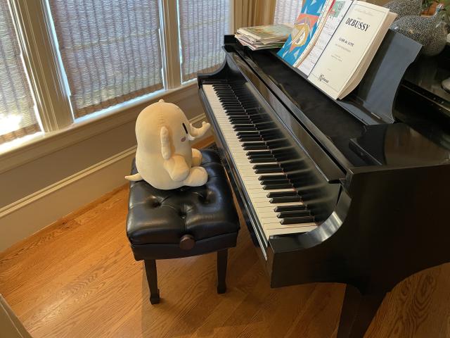 A stuffed Mastodon toy is sitting in front of an open piano. One of the note books reads “Debussy”. 