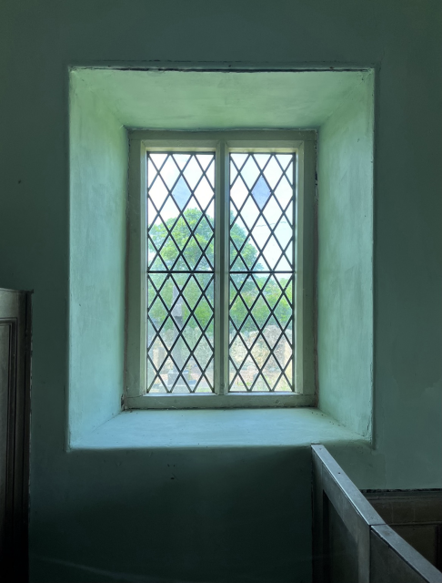 A rectangular window with lattice leading through which can be seen trees, a slate cross, and a stone wall.  The window is recessed into phthalo green walls, the sides of the recess lighter than the facing wall on account of sunlight.  The sides of two wooden box pews are visible to the left of the window and beneath it to the right.
