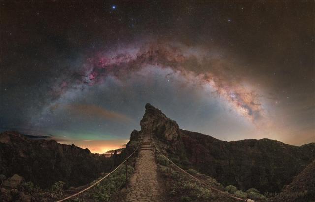 A star filled sky shows the arch of the central band of our Milky Way galaxy across the top of the image. In the foreground is a rocky landscape with a hill ahead and a pathway that leads to stairs up that hill.